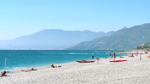 A pebble beach with people sunbathing and swimming, a red boat, and mountains in the background. - Cosy apartment for 3 people with WIFI, TV, balcony and pets allowed (Vallecrosia)