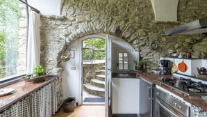 Une cuisine aux murs en pierre, avec un cuisinière à gaz et une vue sur l’extérieur, accessible par un passage en arc de cercle.