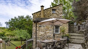 Un bâtiment en pierre doté d’une table et de chaises sur une terrasse, entouré d’une clôture en pierre et de verdure.
