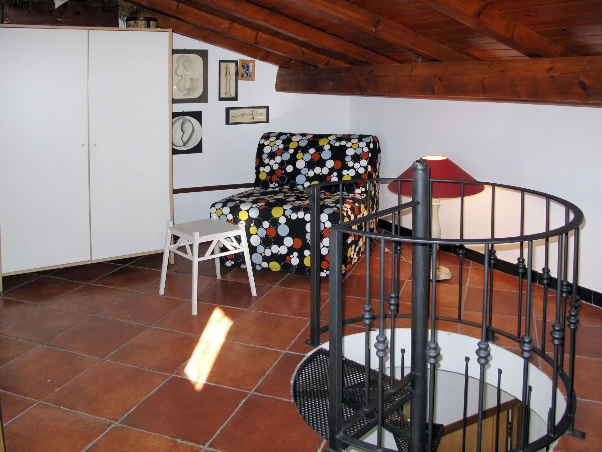 Une pièce avec un meuble blanc, une chaise à pois, un petit tabouret blanc, un escalier en colimaçon et un mur orné de tableaux.