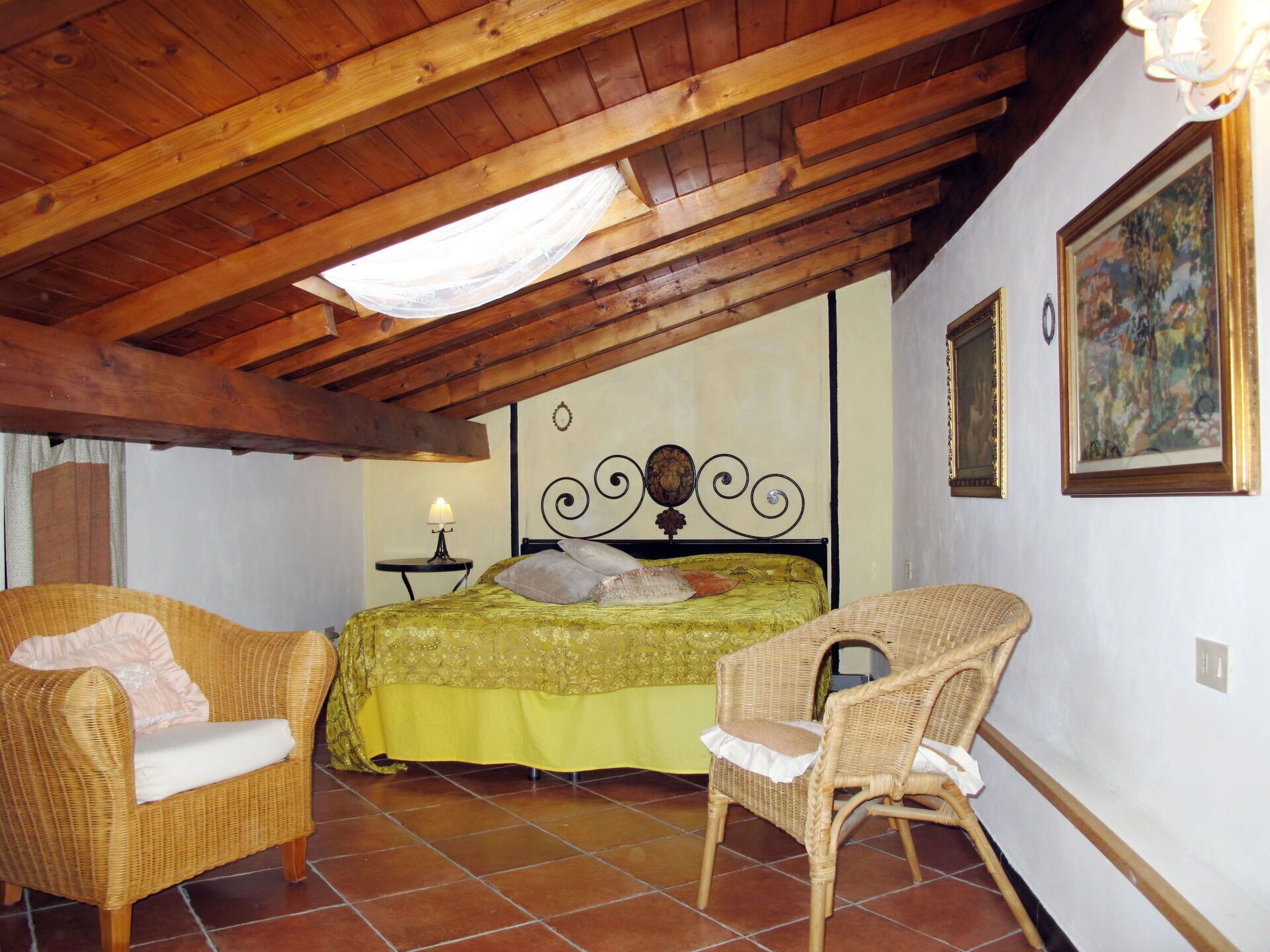 A cozy room with a bed, two wicker chairs, a small table with a lamp, and a painting on the wall.