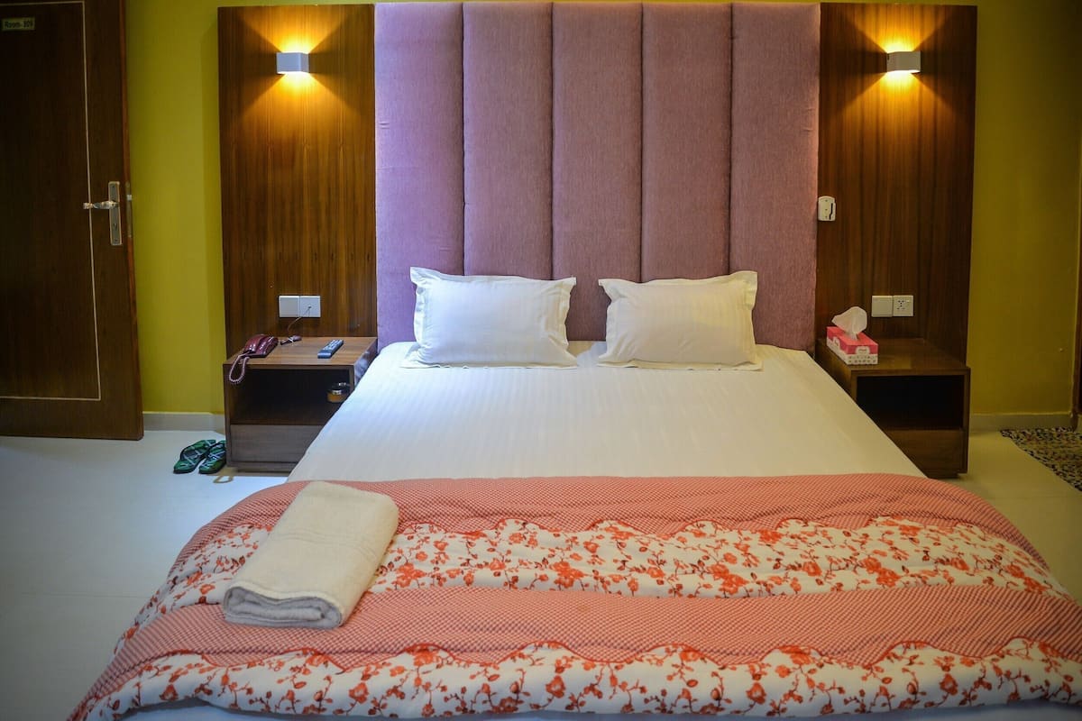 Una habitación de hotel con una cama grande, dos mesitas de noche, un cabecero de madera y una mesita con una lámpara.