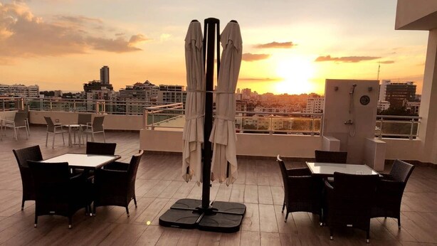 A rooftop terrace with two tables and chairs, a central umbrella stand, and a cityscape view during sunset. - Santo Domingo Apartment with Rooftop Pool with Ocean & City Views (Santo Domingo)