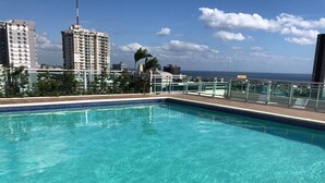 A rooftop pool with clear blue water, surrounded by a metal railing and palm trees. - Santo Domingo Apartment with Rooftop Pool with Ocean & City Views (Santo Domingo)