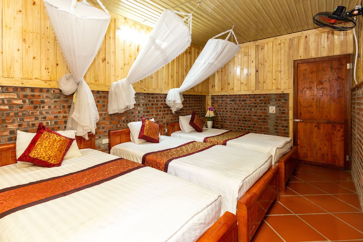 A room with two beds, each with white linens and red and gold pillows, a brick wall, and a wooden door.