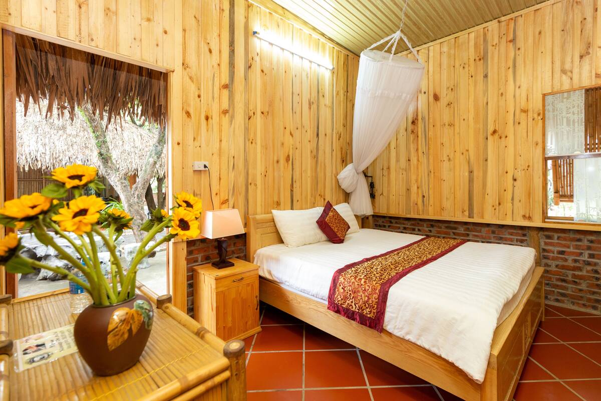 A wooden room with a bed, a nightstand, a vase of sunflowers, and a window with a view of trees.