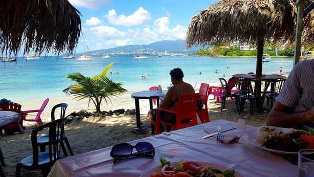 Een restaurant aan het strand met terras, uitzicht op zee en boten op de achtergrond.
