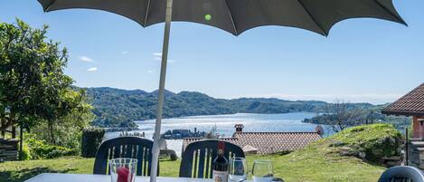 An outdoor table with a large umbrella, a bottle of wine, and glasses, overlooking a scenic lake and hills.