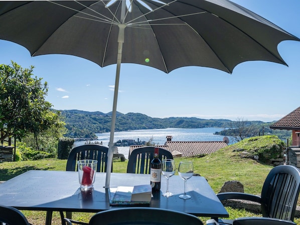 An outdoor table with a large umbrella, a bottle of wine, and glasses, overlooking a scenic lake and hills.