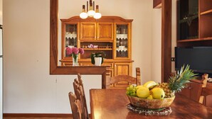 Une table à manger en bois, entourée de chaises, avec un saladier de fruits dessus, et, au fond, un buffet en bois doté de portes en verre.