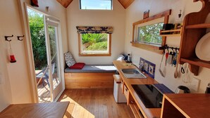 Cozinha compacta com piso de madeira, um banco embutido com almofada vermelha, uma janela com vista para o verde e uma pia com diversos utensílios.