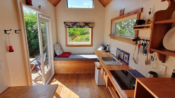 Cocina compacta con piso de madera, un banco empotrado con un cojín rojo, una ventana con vistas a follaje y un fregadero con diversos utensilios.
