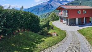 A red house with a balcony, surrounded by a gravel driveway and lush greenery, with mountains in the background. - Beautiful private villa for 8 guests with WIFI, TV, terrace and pets allowed (Peglio)
