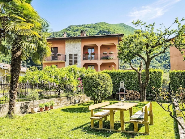 A two-story building with balconies, a garden with a wooden table and benches, and a mountain in the background.