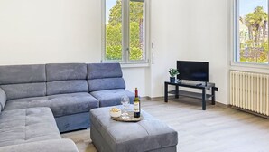 Ett modernt vardagsrum med en grå hörnsoffa, ett litet soffbord med en flaska och glas, en tv på ett tv-bord och stora fönster med utsikt över träd och byggnader.