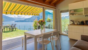 Ein modernes Zimmer mit einer großen Schiebeglastür, die auf einen Balkon mit Blick auf die Berge führt. Im Inneren befindet sich ein weißer Tisch mit Stühlen, eine Blumenvase sowie eine Küchenzeile mit Schränken und Geräten.