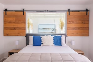 Une chambre Ă coucher avec un lit, deux tables de chevet, une vue sur la mer par une fenĂȘtre et des panneaux muraux en bois.