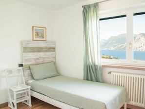 A single bed with a wooden headboard, a small white chair, a radiator, and a window with a view of a mountainous coastline. - Stunning  villa for 6 people with A/C, WIFI, TV, terrace and pets allowed (Malcesine)
