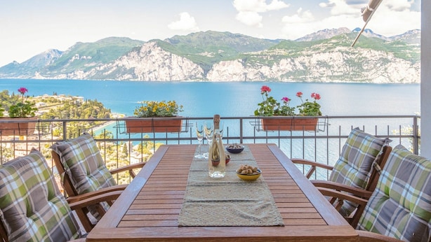 Una mesa de madera con un mantel individual, dos macetas y una botella de agua en un balcón con vista al mar y montañas al fondo.