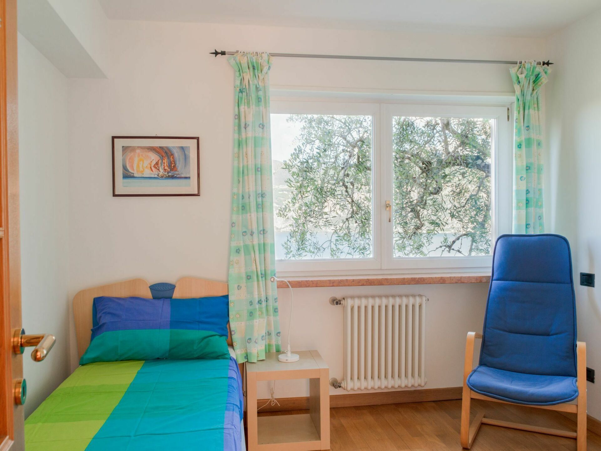 A bedroom with a bed, a chair, a radiator, a window with curtains, and a painting on the wall.