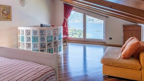 Una habitación con techo de madera, una cama con colcha a rayas, una mesita con superficie de vidrio, un sofá con cojines y una ventana con vistas a un lago y montañas.