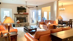 A spacious living room with a stone fireplace, leather sofas, a glass coffee table, and a dining area with a chandelier. - Lake Front Home. 3 Large Living Spaces. Screened Porches. Pet Friendly. (Lavonia)
