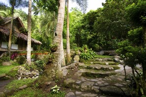 Ein traditionelles Haus mit einem Reetdach, umgeben von üppiger Vegetation und einem Steinelement für Wasser.