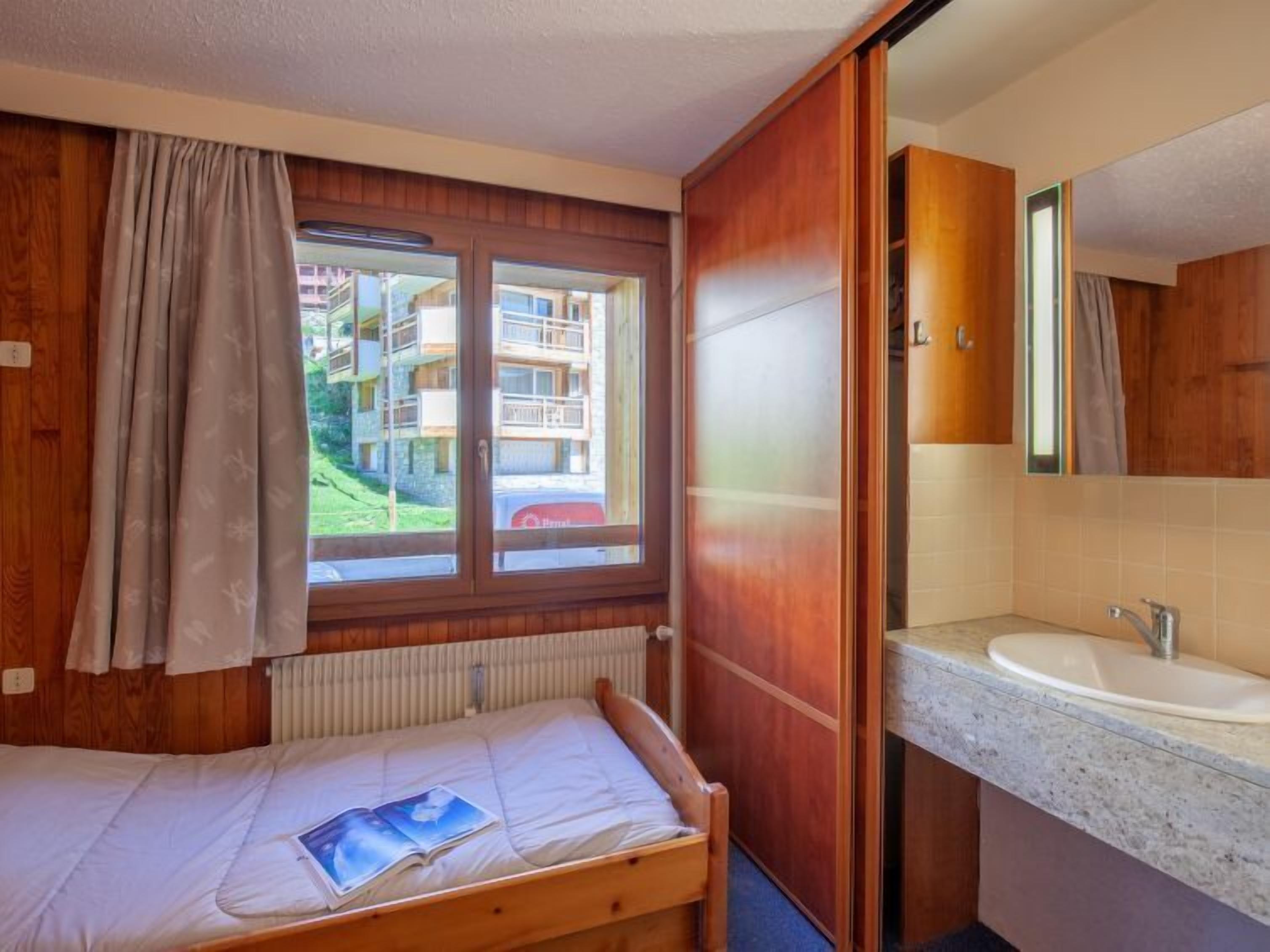 A small, single-bed room with a window, a wooden wardrobe, and a bathroom with a sink and mirror.