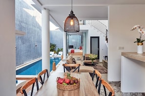 A modern dining area with a pool view, a wooden table with fruit, and a hanging light fixture. - 🏖 Private room with pool, 5min walk to Echo Beach (Seminyak)