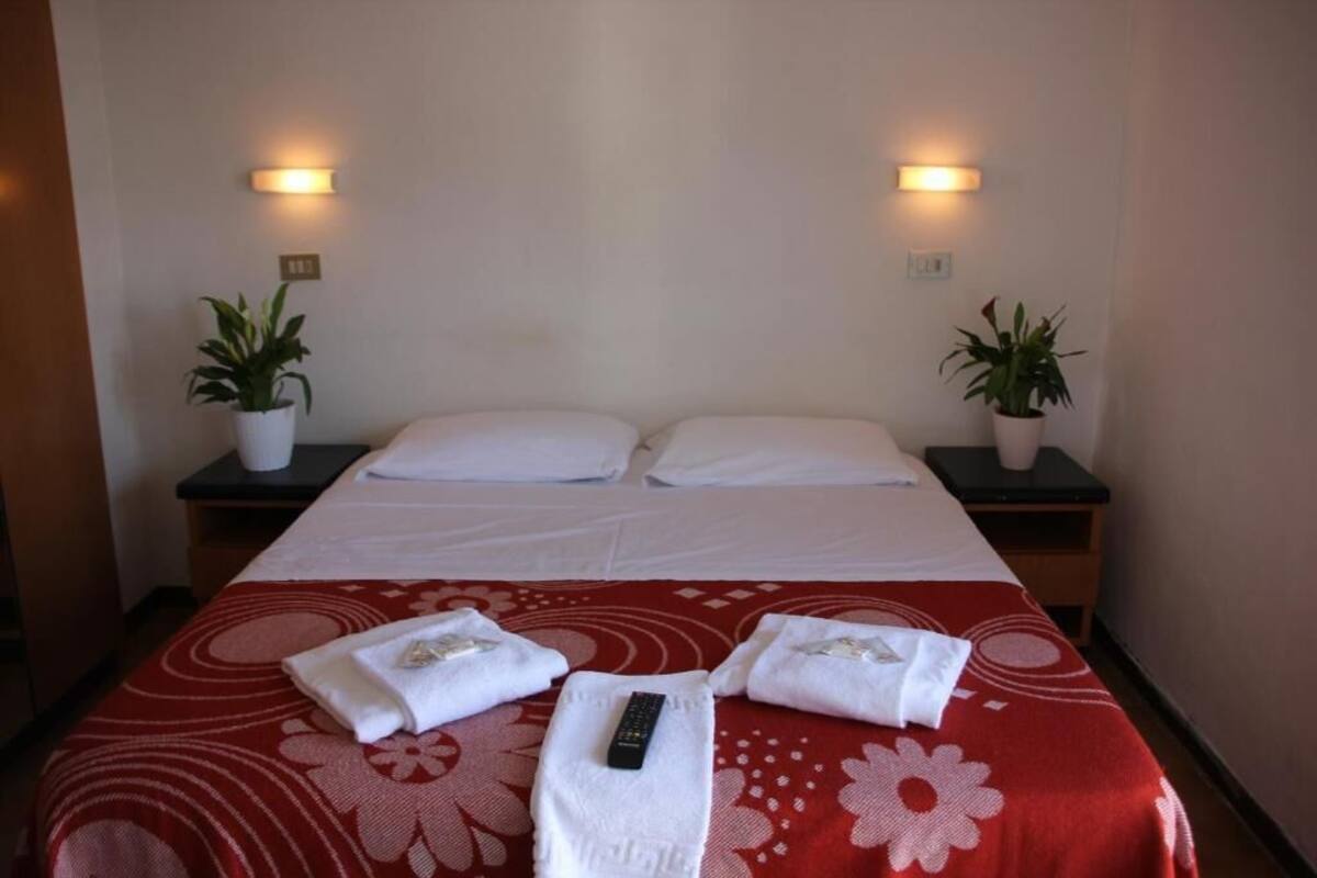 A hotel room with a bed, two bedside tables with potted plants, and a red floral bedspread.