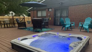 Een hottub op een houten terras met tuinmeubilair en een bakstenen gebouw op de achtergrond.