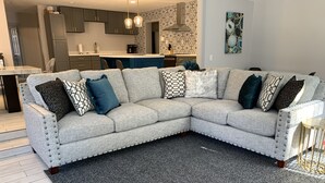 Een moderne woonkamer met een grijze hoekbank, een donkergrijze vloerkleed en een keuken op de achtergrond.