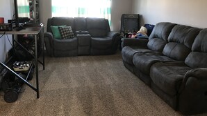 Een woonkamer met een donker leren bank, een fauteuil, een tafeltje met elektronische apparaten en een raam met gordijnen.