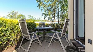 Un patio exterior con una mesa y dos sillas, una puerta a la derecha y vistas a árboles y arbustos.