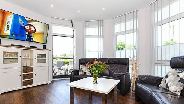 Una sala de estar moderna con un televisor de pantalla plana, una mesa de centro blanca, un sofá de cuero negro y un comedor con mesa y sillas.
