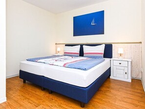 A neatly made bed with a blue headboard, a bedside table with a lamp, and a wall-mounted painting of a sailboat.