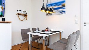 Ein Essbereich mit einem weißen Tisch, vier grauen Stühlen, schwarzen Pendelleuchten und einem Seestück an der Wand.