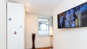 Ein Flur mit Holzboden, einer weißen Tür mit Ankerverzierungen, einem Fenster mit Jalousien, einer Stehlampe und einem an der Wand montierten Fernseher, der eine Szene zeigt.