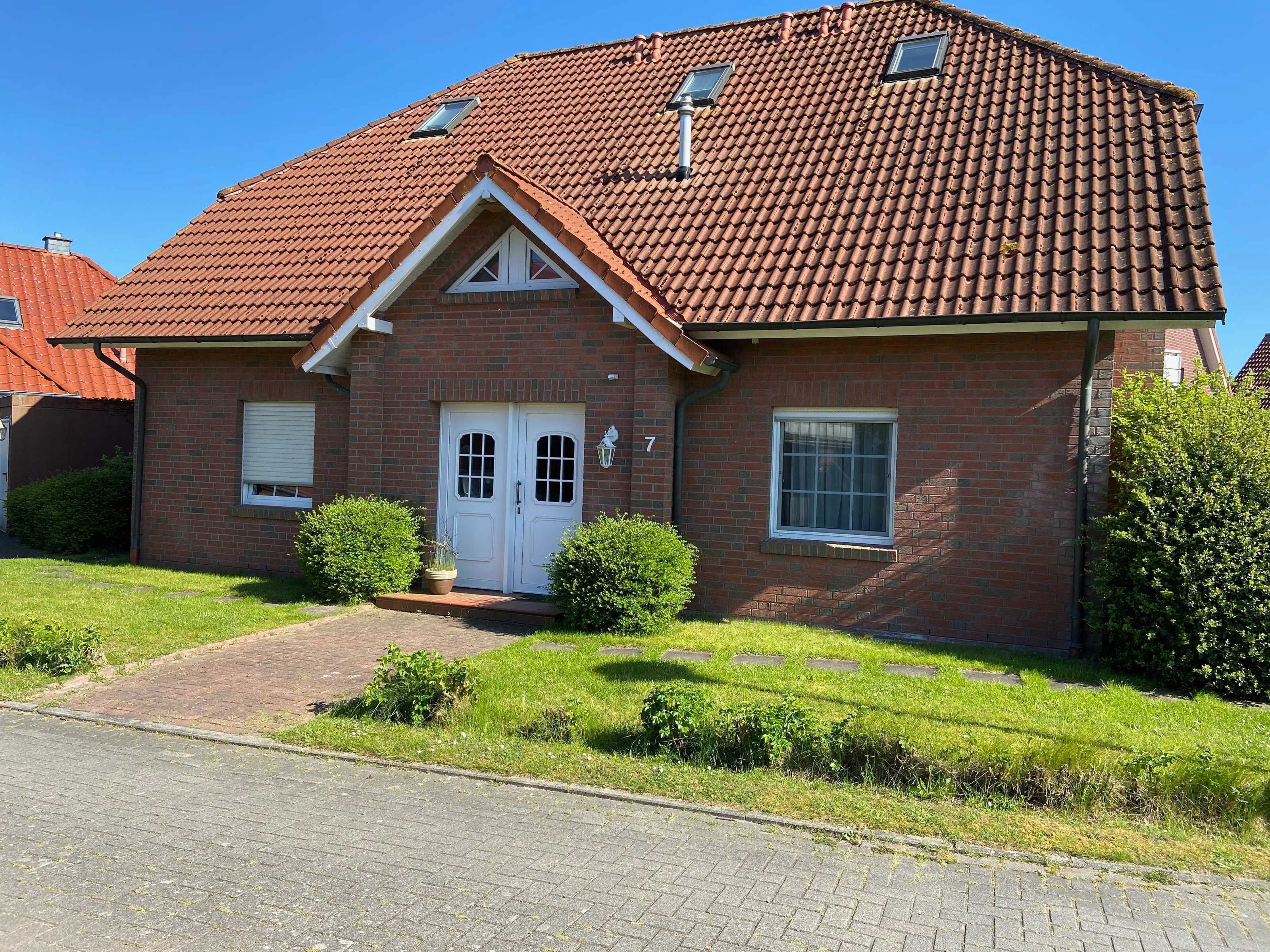 Ein zweistöckiges Backsteinhaus mit rotem Ziegeldach, einem Hauseingang mit weißer Tür und einem kleinen Garten mit Büschen und einer Kübelpflanze.