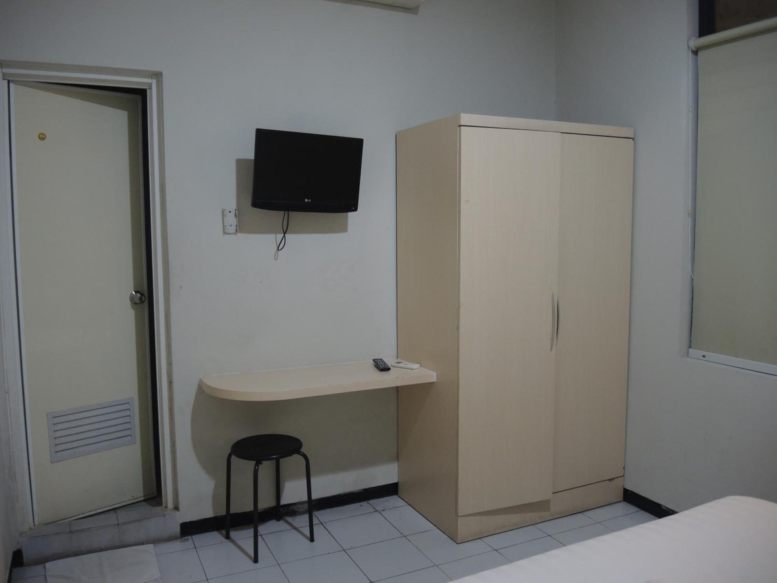 A small, simple room with a bed, a wardrobe, a desk with a chair, and a television mounted on the wall.
