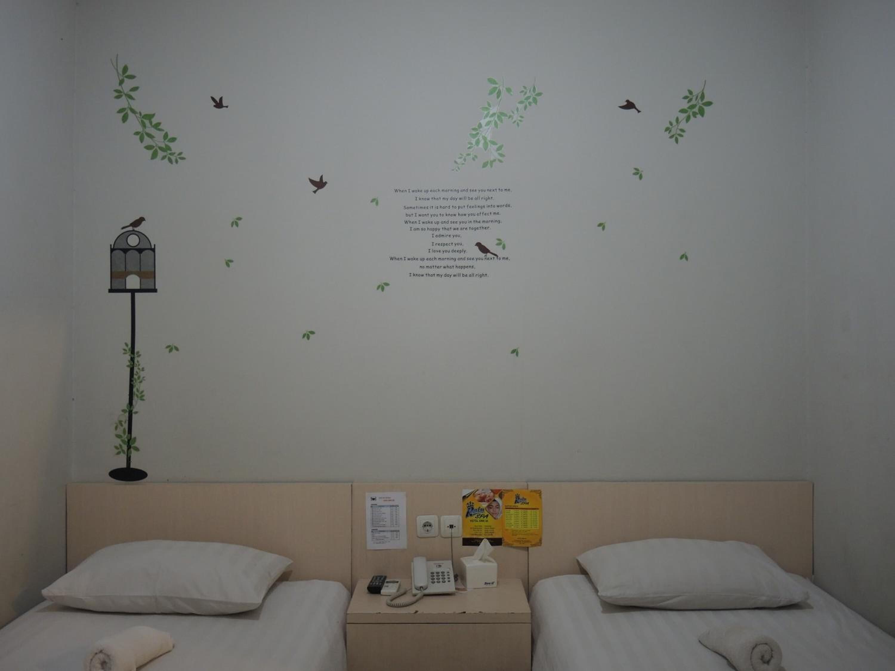 Habitación de hotel con dos camas, una mesita de noche con teléfono y pañuelos, y un adhesivo mural con pájaros y hojas.