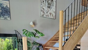 Un escalier en bois, avec une paroi de verre à sa base, un pot de fleurs et des œuvres d’art accrochées aux murs.