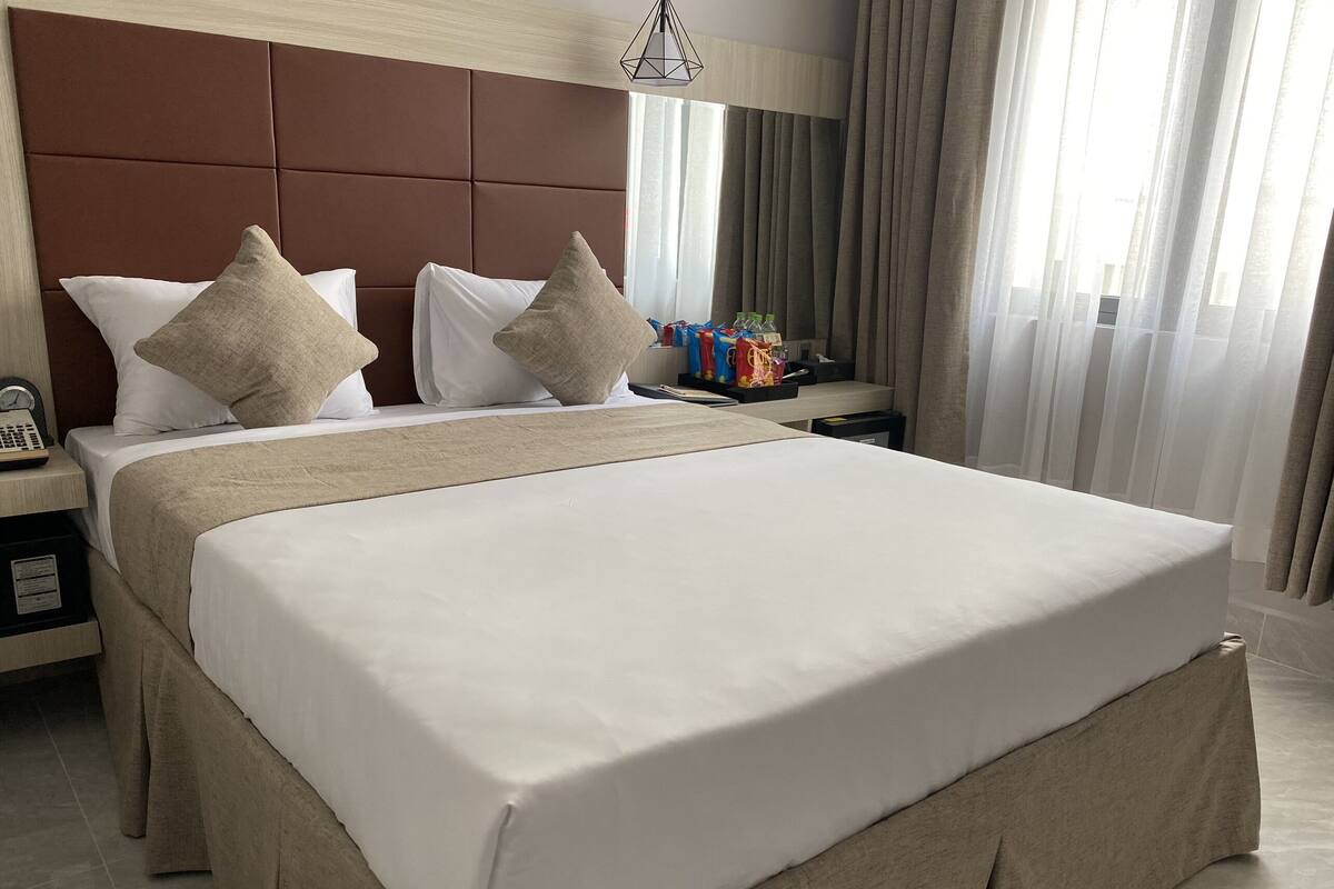 Hotellihuone, jossa on suuri sänky, pääty, yöpöytä lampun kanssa ja ikkuna, jossa on verhot.