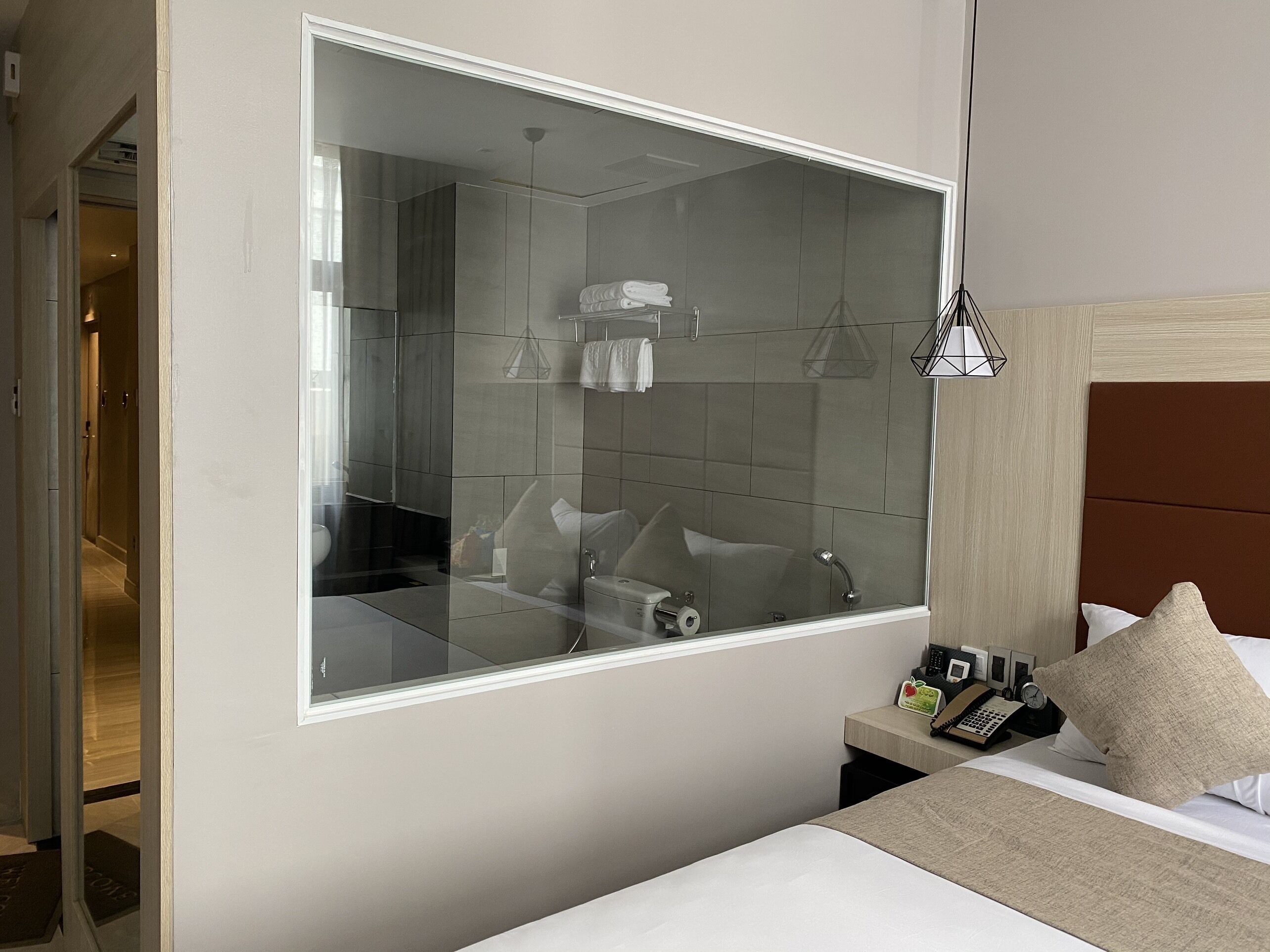 Une chambre d’hôtel moderne avec un grand miroir, un lit doté d’une tête de lit, une table de chevet avec un téléphone et une horloge, et une salle de bain visible à travers le miroir.