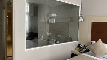 A modern hotel room with a large mirror, a bed with a headboard, a bedside table with a phone and a clock, and a bathroom visible through the mirror.