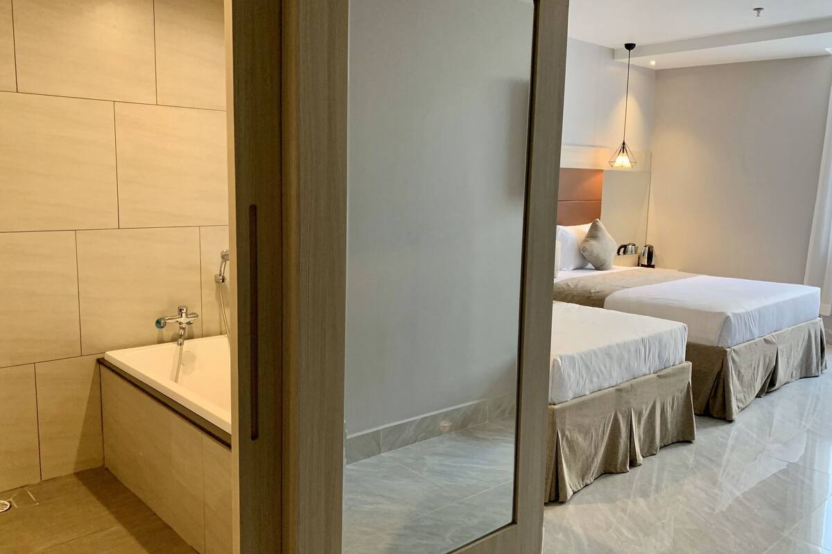 Moderni hotellihuone, jossa on suuri peili, kylpyamme, kaksi sänkyä ja pieni pöytä, jonka päällä on vedenkeitin.