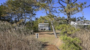A wooden pathway leading to a two-story house with a balcony, surrounded by pine trees and dry grass.
