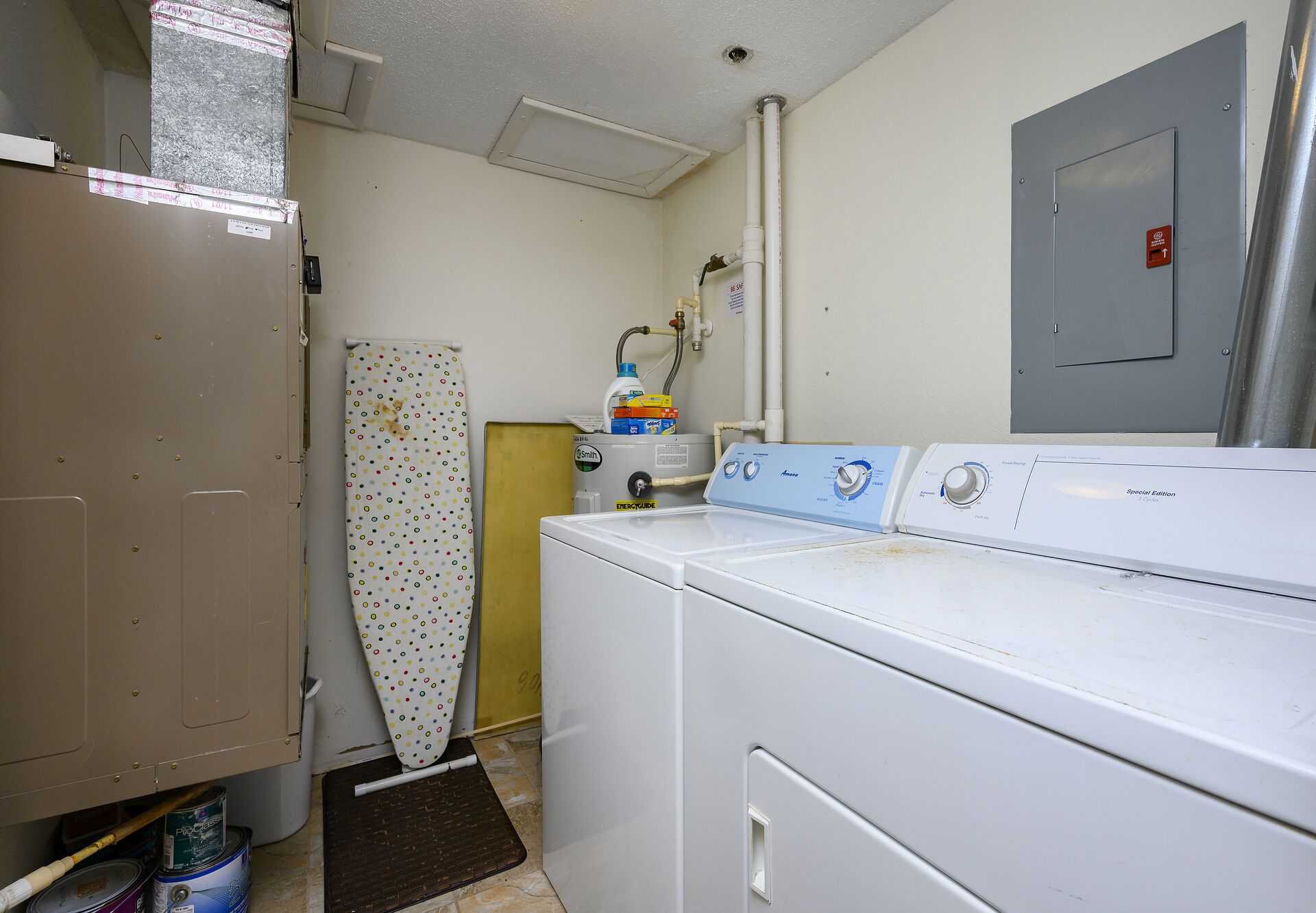 Une buanderie équipée d’un lave-linge, d’un sèche-linge et d’une pièce technique contenant un chauffe-eau et une porte dotée d’un détecteur d’incendie.