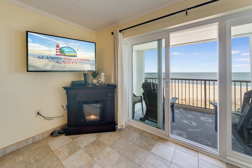 A living room with a fireplace, a television displaying 'SEASIDE VACATIONS & SALES', and a large sliding glass door leading to a balcony with beach views.