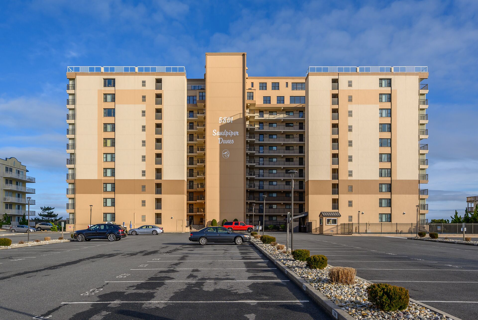 A multi-story residential building with a central entrance, parking lot, and a clear sky.